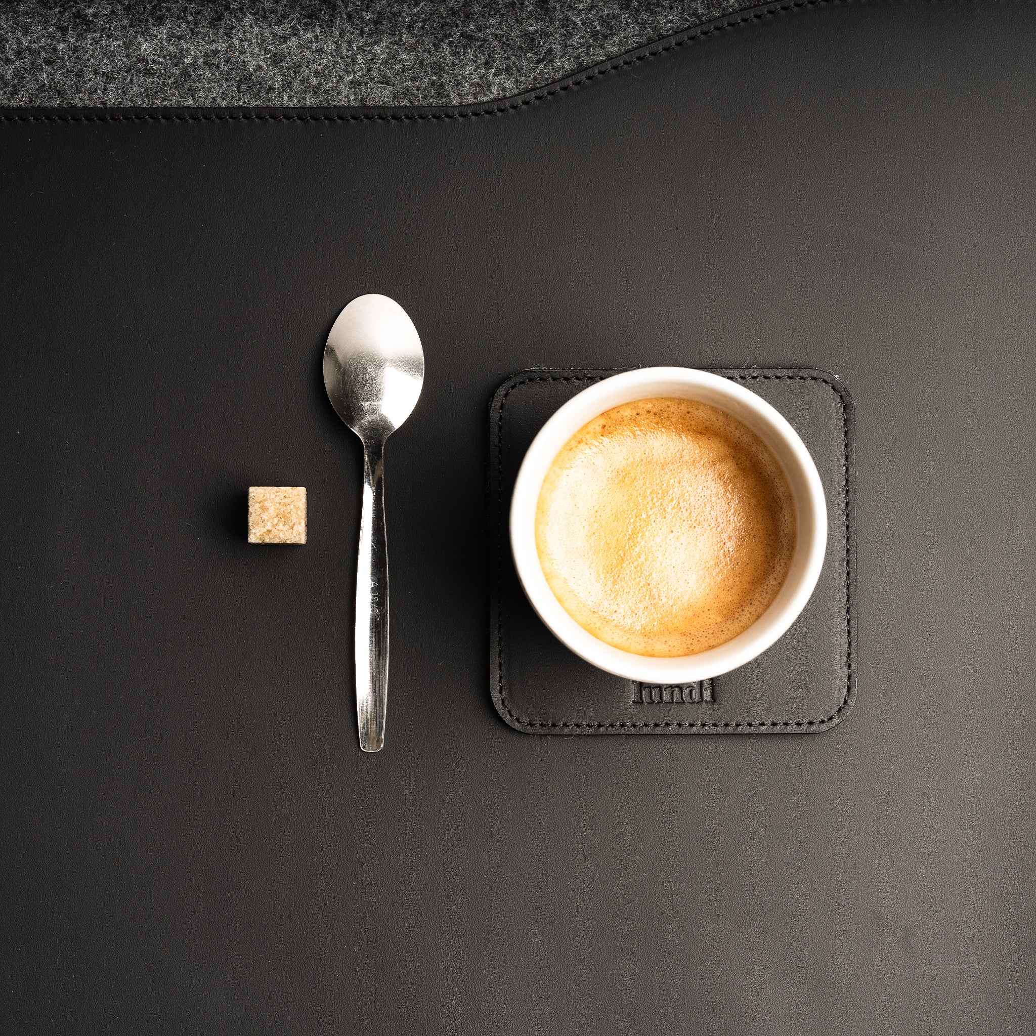 Sous tasse en cuir noir avec tasse de café, cuillère et sucre posés sur un bureau élégant.