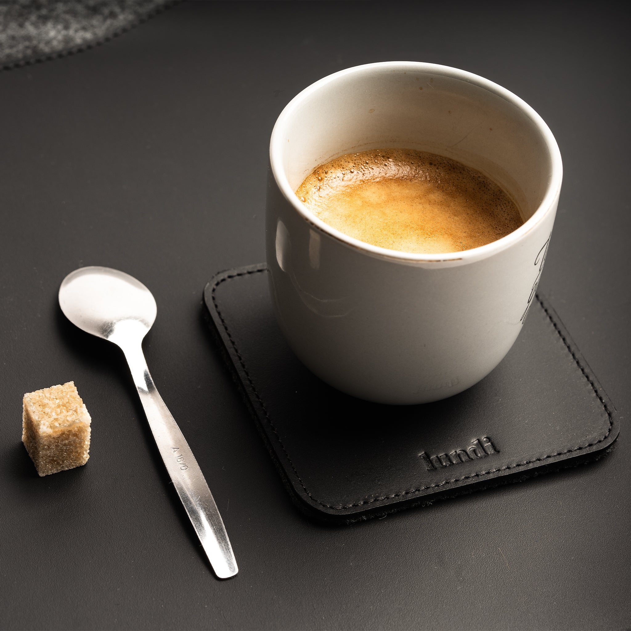 Sous verre noir en cuir avec tasse blanche, sucre et cuillère en métal sur surface noire.