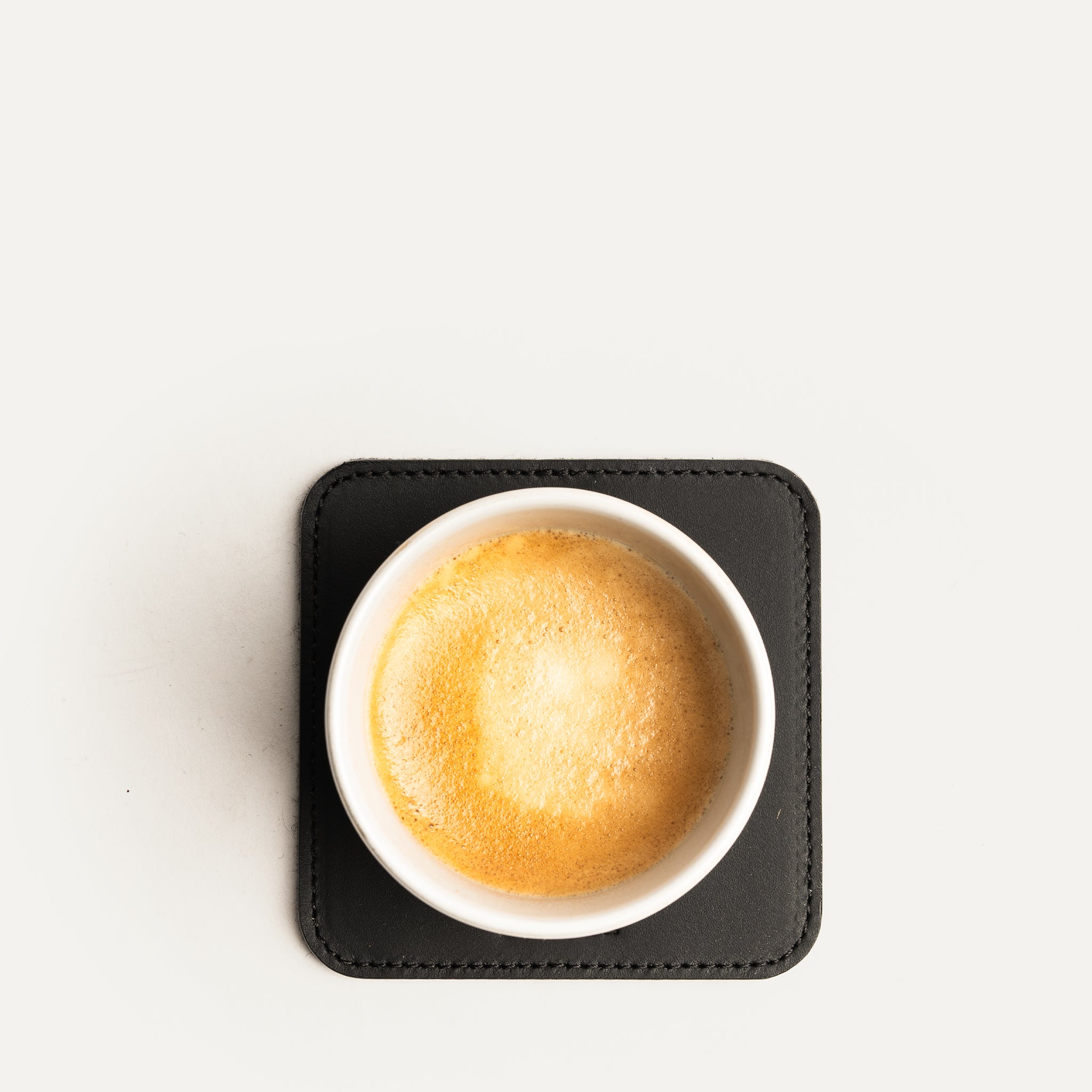 Dessous de verre noir en cuir avec tasse de café vue du dessus, posé sur fond blanc minimaliste.