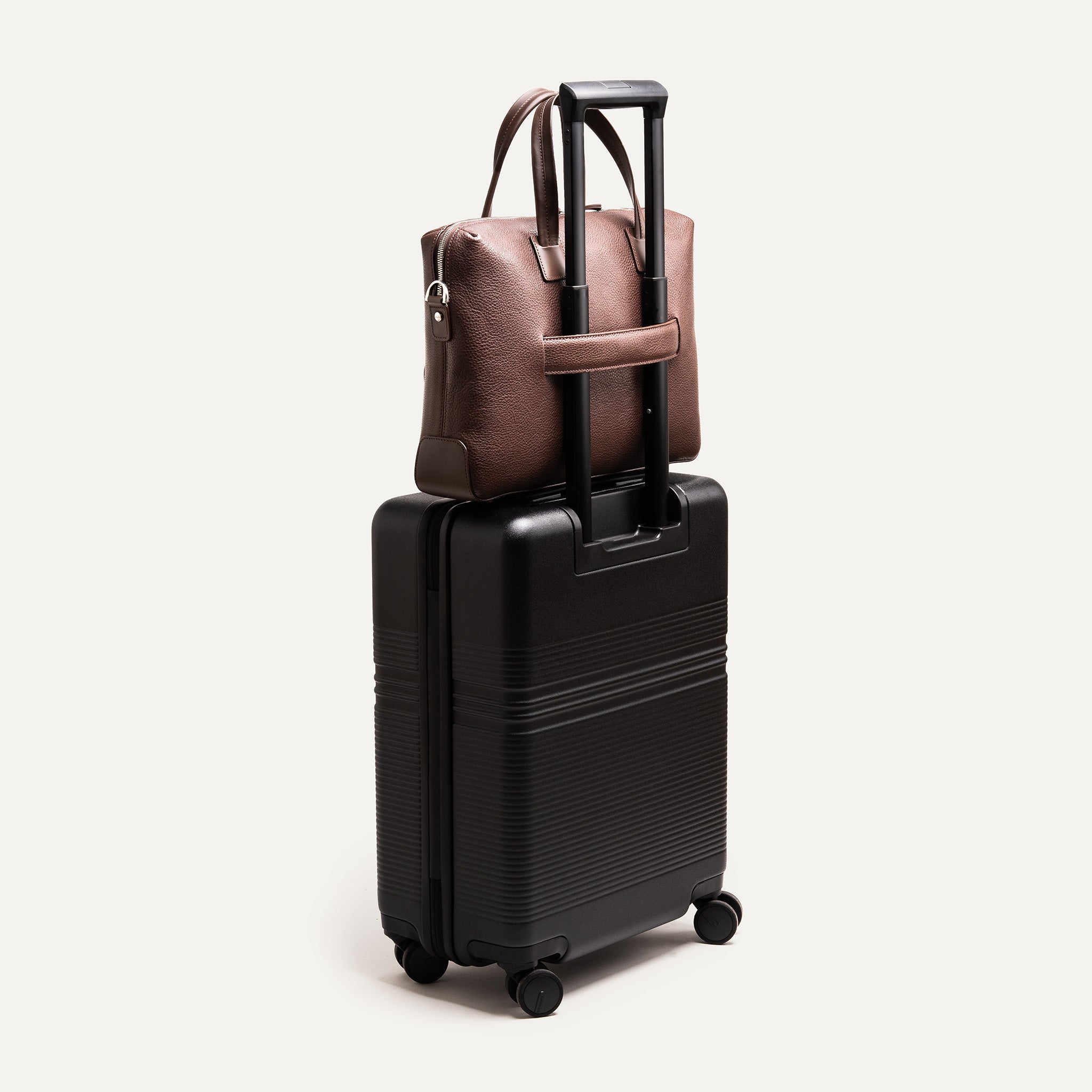 Sac à bandoulière homme en cuir marron accroché à la poignée d’une valise trolley noire, idéal pour les voyages d'affaires.