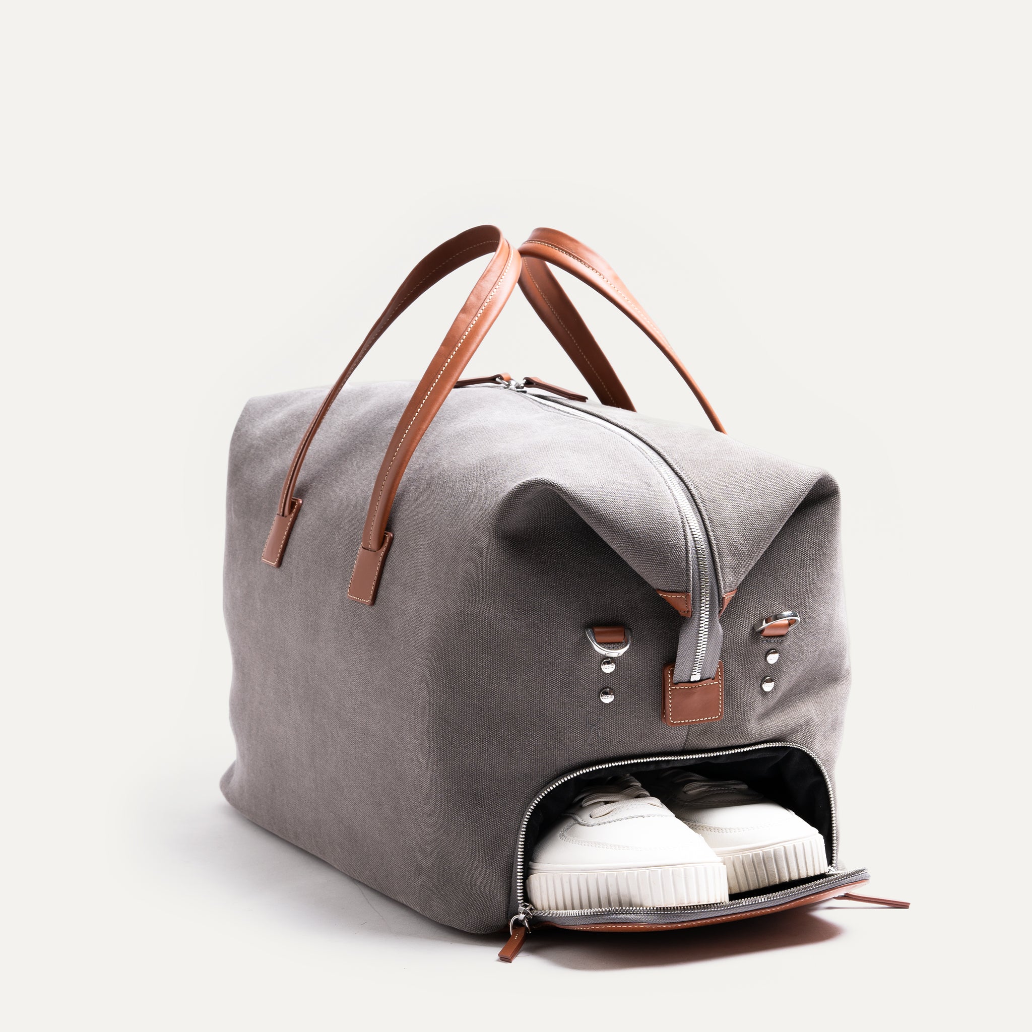 Men’s leather travel bag angled to reveal the side shoe compartment unzipped, displaying white sneakers tucked inside.