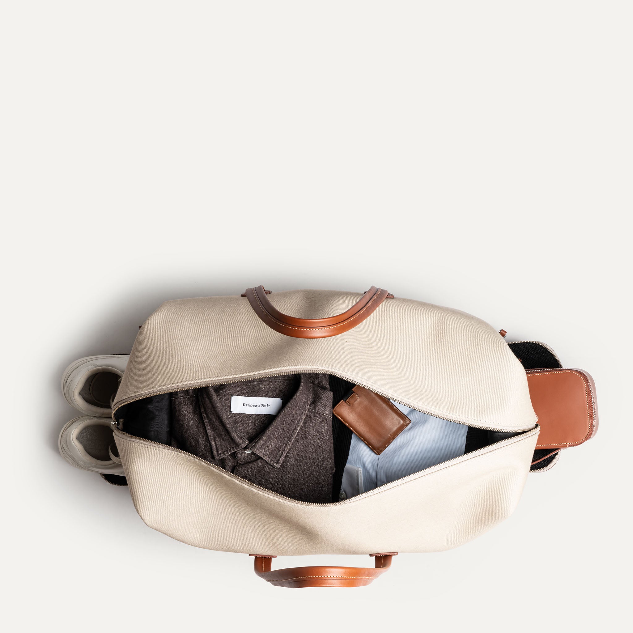 Vue intérieure d’un sac week-end beige en cuir et toile contenant chemises, portefeuille et paire de sneakers.