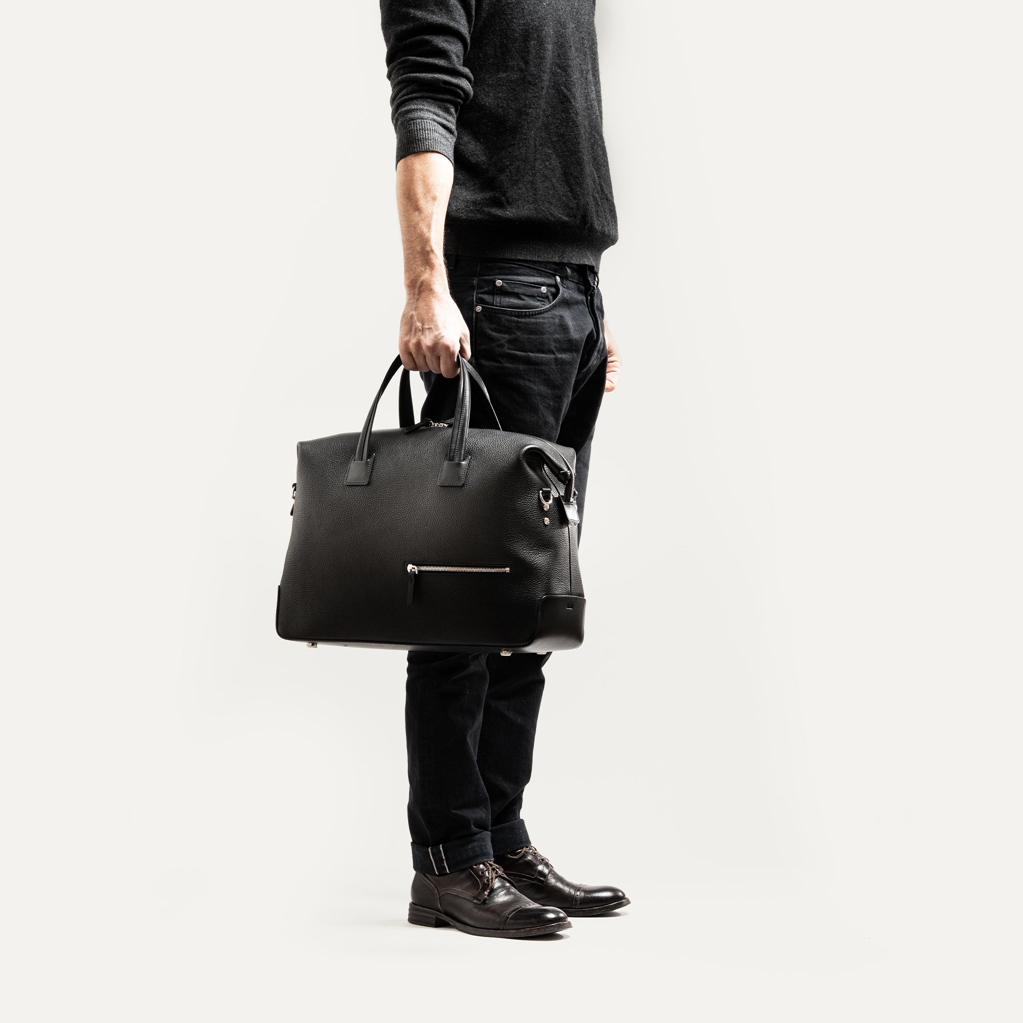Sac de voyage homme en cuir noir, format 48 heures, porté à la main pour déplacement professionnel élégant.